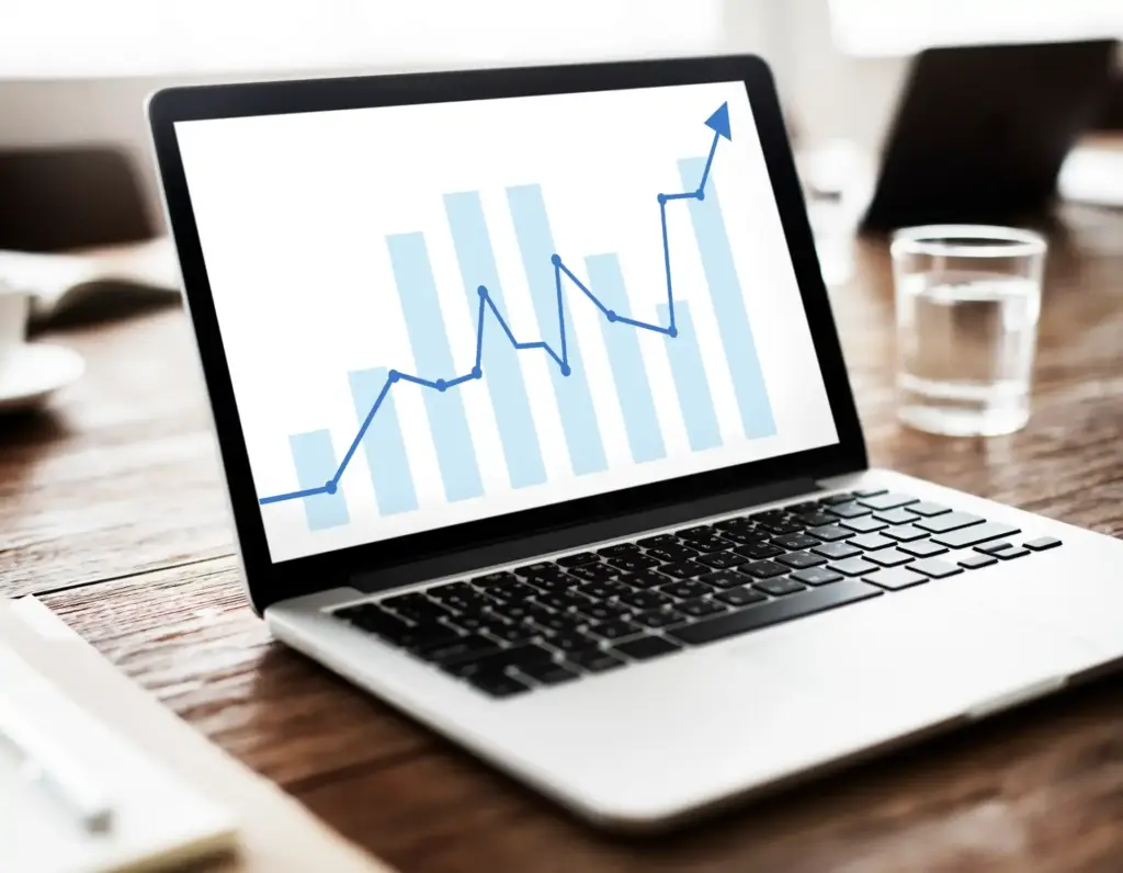 Notebook em uma mesa com gráfico de crescimento em azul na tela, simulando análise de dados e indicadores de desempenho, com setas subindo e barras no fundo