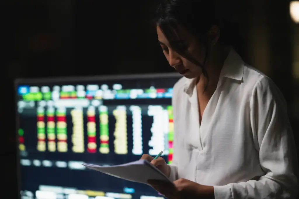 Mulher analisando dados financeiros em escritório com tela de computador exibindo gráficos e números, focada em análise de mercado.