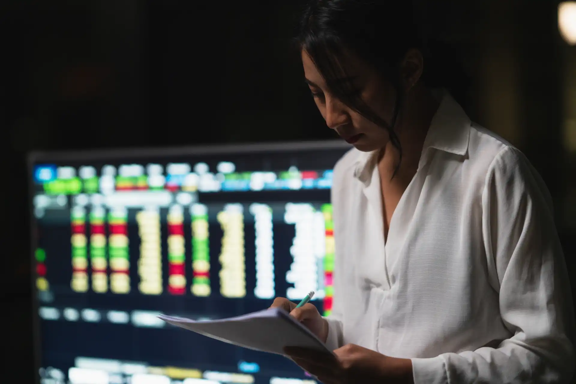 Mulher analisando dados financeiros em escritório com tela de computador exibindo gráficos e números, focada em análise de mercado.