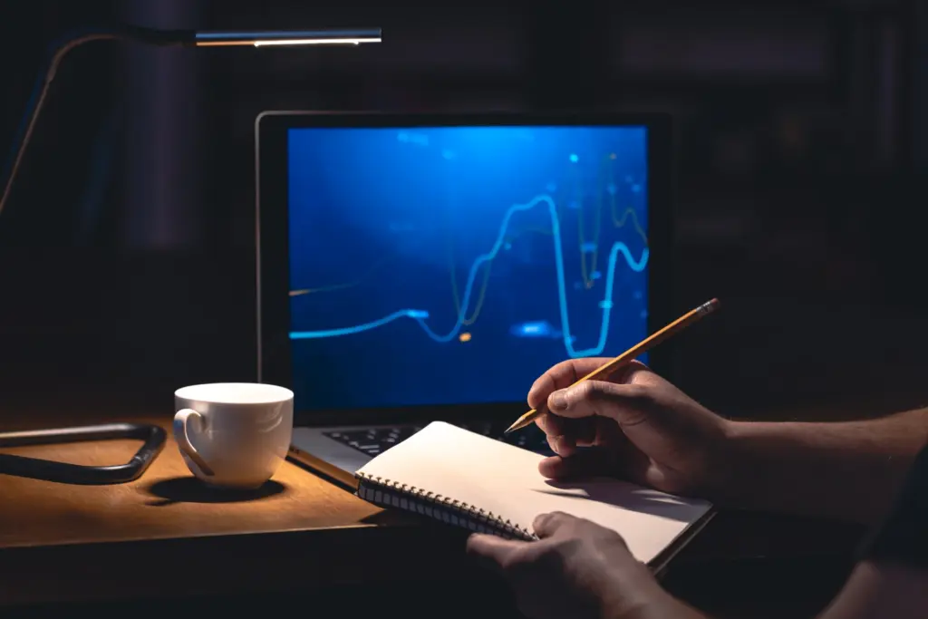 Pessoa anotando ideias em um caderno enquanto observa gráficos de dados em uma tela de computador, ambiente de trabalho com foco em análise de dados e investimentos.