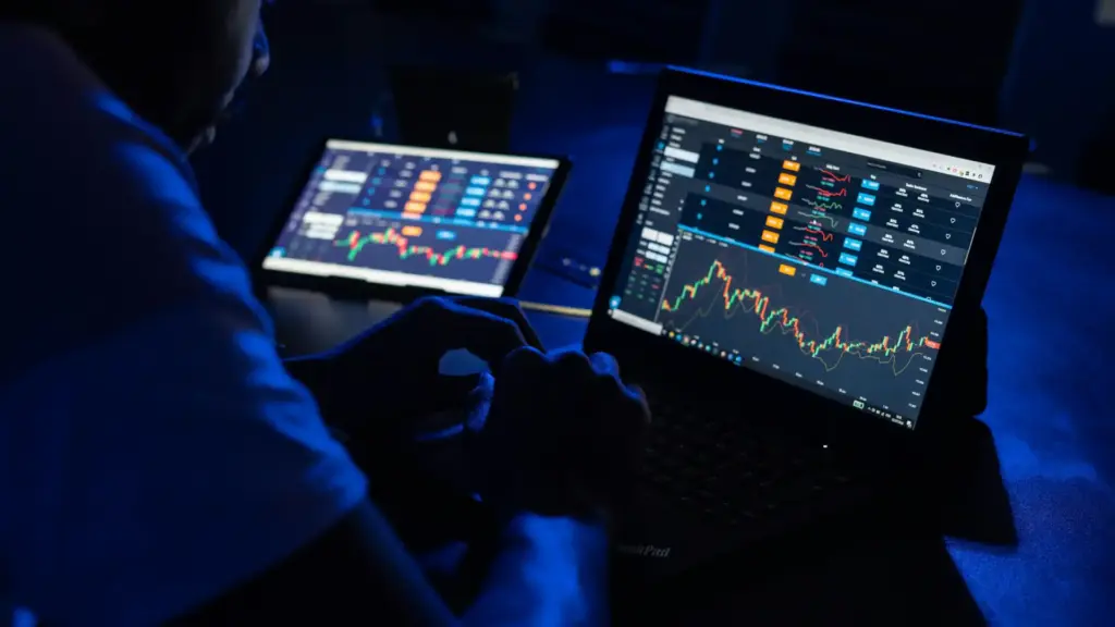 Pessoa analisando gráficos de ações em dois computadores, monitorando o mercado financeiro e realizando operações de trading no ambiente digital.
