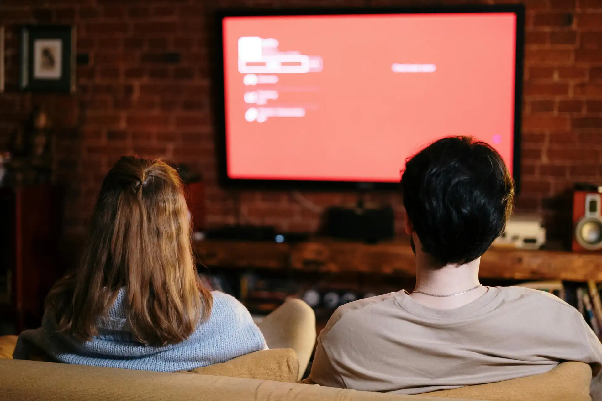 Casal assistindo a televisão em uma sala com decoração moderna de paredes de tijolos, concentrados na tela vermelha exibindo um menu ou playlist.