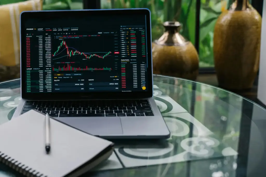 Imagem de um laptop exibindo gráficos de ações na bolsa de valores, acompanhado de uma caneta e caderno, em um ambiente com plantas e vasos decorativos.