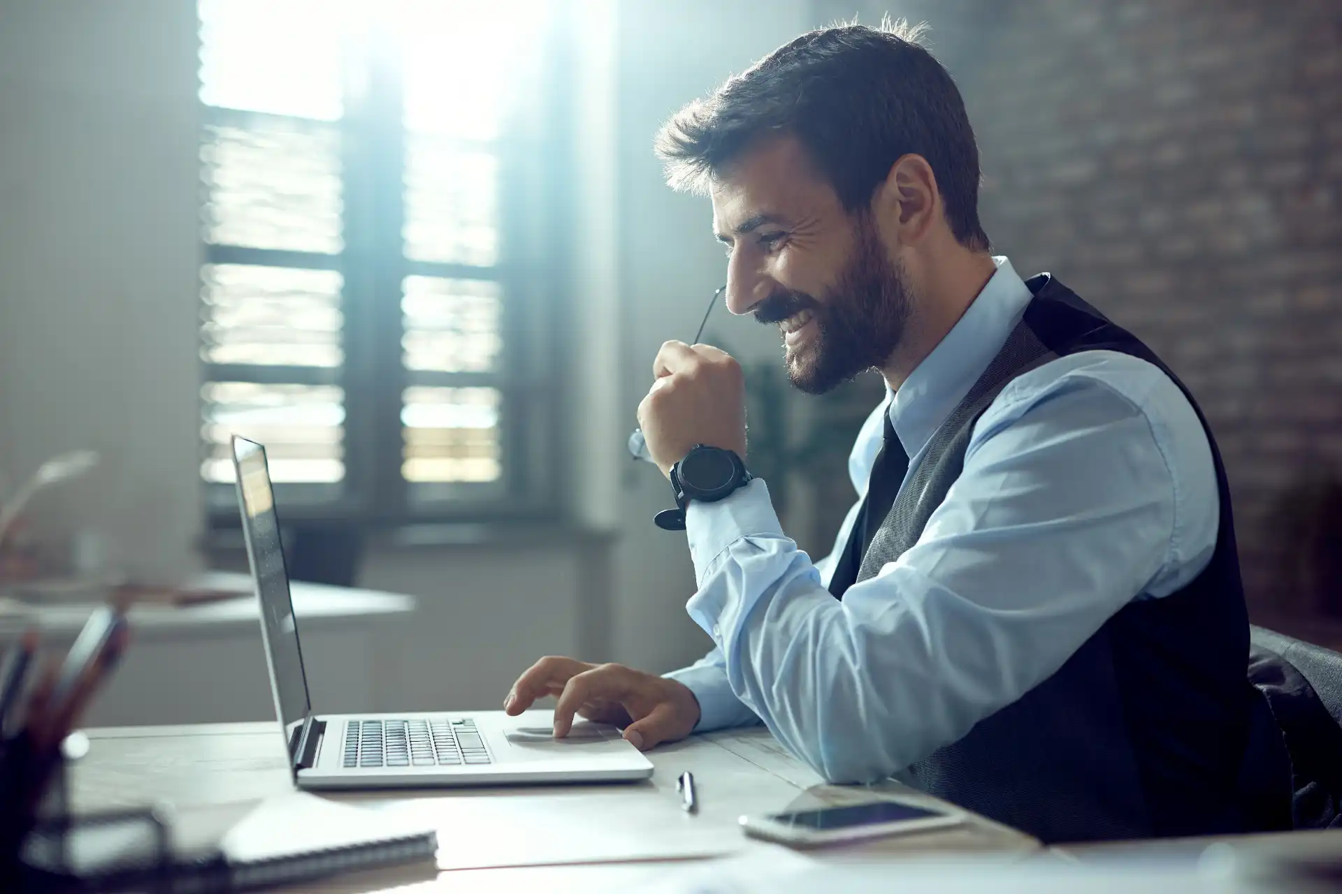 Homem de negócios sorrindo enquanto trabalha com laptop em escritório moderno, simbolizando produtividade e sucesso profissional.
