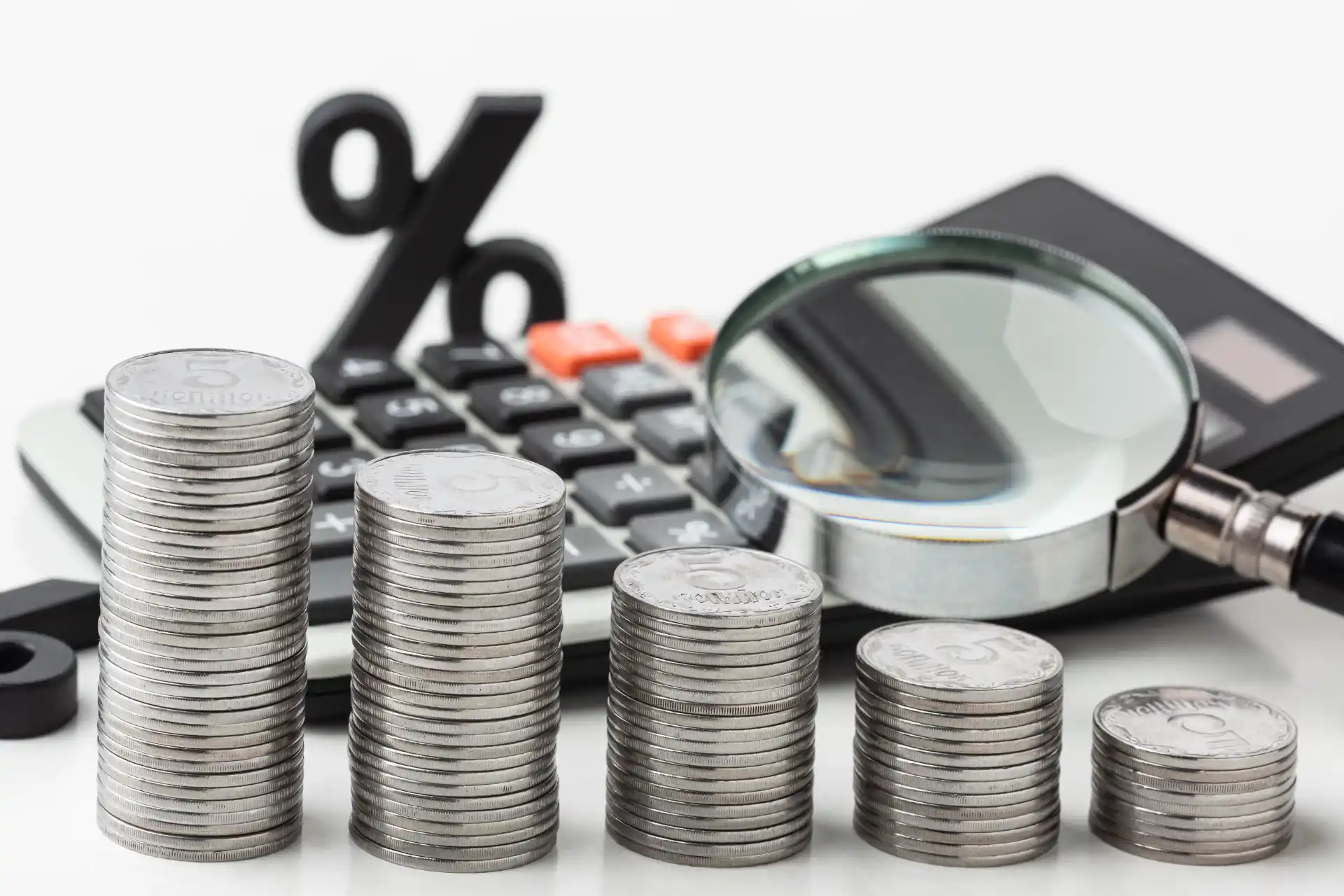 Pilha de moedas de diferentes alturas sobre teclado e lupa de aumento, com símbolo de porcentagem ao fundo, ilustrando economia, análise financeira e redução de custos.